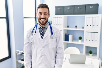 Wall Mural - Handsome hispanic man wearing doctor uniform and stethoscope at medical clinic looking positive and happy standing and smiling with a confident smile showing teeth