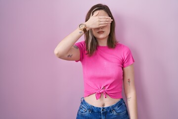 Poster - Blonde caucasian woman standing over pink background covering eyes with hand, looking serious and sad. sightless, hiding and rejection concept