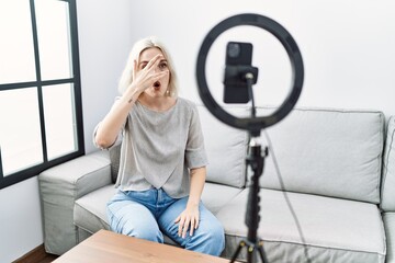 Poster - Young caucasian woman recording vlog tutorial with smartphone at home peeking in shock covering face and eyes with hand, looking through fingers with embarrassed expression.