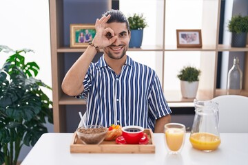 Wall Mural - Hispanic man with long hair sitting on the table having breakfast smiling happy doing ok sign with hand on eye looking through fingers