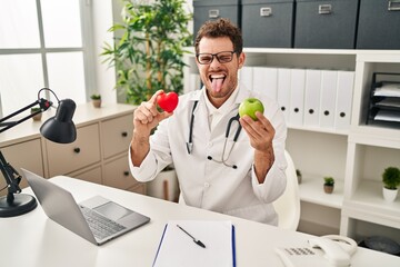 Canvas Print - Young hispanic man working at dietitian clinic sticking tongue out happy with funny expression.