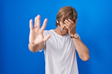 Poster - Middle age man standing over blue background covering eyes with hands and doing stop gesture with sad and fear expression. embarrassed and negative concept.