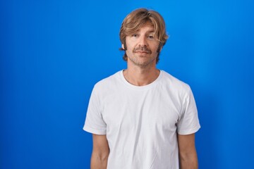 Poster - Middle age man standing over blue background relaxed with serious expression on face. simple and natural looking at the camera.