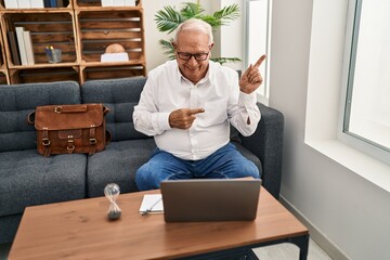 Canvas Print - Senior therapist with grey hair doing online session at consultation office smiling and looking at the camera pointing with two hands and fingers to the side.