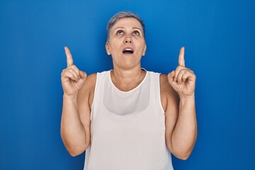 Canvas Print - Middle age caucasian woman standing over blue background amazed and surprised looking up and pointing with fingers and raised arms.