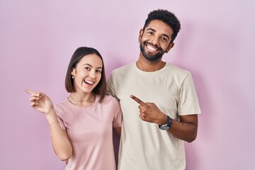 Canvas Print - Young hispanic couple together over pink background with a big smile on face, pointing with hand finger to the side looking at the camera.