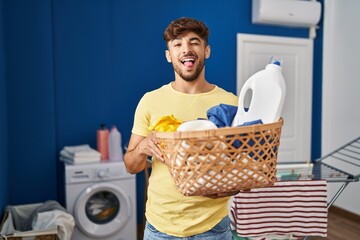 Poster - Arab man with beard holding laundry basket and detergent bottle sticking tongue out happy with funny expression.