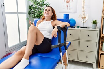 Sticker - Young redhead woman lying on rehabilitation bed holding crutches angry and mad screaming frustrated and furious, shouting with anger. rage and aggressive concept.