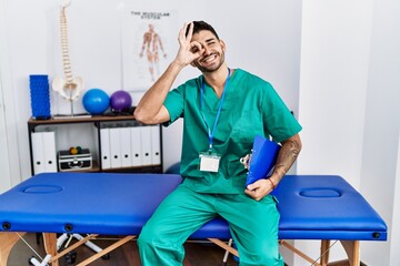 Wall Mural - Young physiotherapist man working at pain recovery clinic doing ok gesture with hand smiling, eye looking through fingers with happy face.