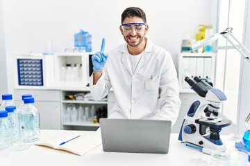 Canvas Print - Young hispanic man working at scientist laboratory with laptop smiling with an idea or question pointing finger with happy face, number one