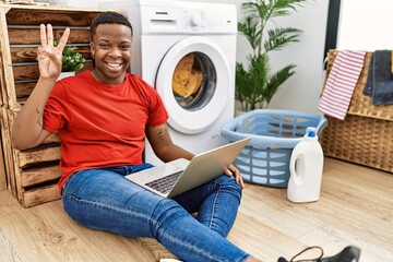 Canvas Print - Young african man doing laundry and using computer showing and pointing up with fingers number three while smiling confident and happy.