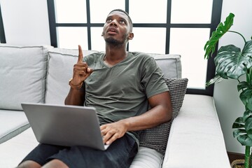 Poster - Young african american man using laptop at home sitting on the sofa amazed and surprised looking up and pointing with fingers and raised arms.