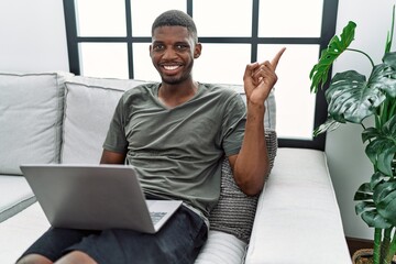 Wall Mural - Young african american man using laptop at home sitting on the sofa with a big smile on face, pointing with hand finger to the side looking at the camera.