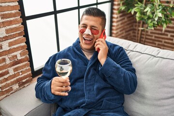 Poster - Hispanic young man wearing bathrobe and eye bags patches drinking wine speaking on the phone winking looking at the camera with sexy expression, cheerful and happy face.