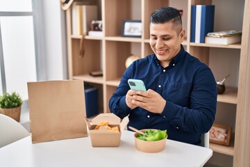 Wall Mural - Hispanic young man eating take away food using smartphone smiling with a happy and cool smile on face. showing teeth.