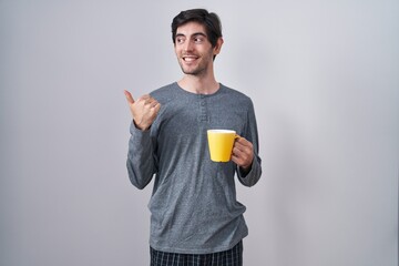 Poster - Young hispanic man wearing pajama drinking a cup of coffee smiling with happy face looking and pointing to the side with thumb up.