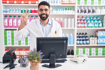 Sticker - Hispanic man with beard working at pharmacy drugstore showing and pointing up with fingers number three while smiling confident and happy.