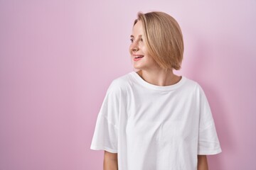 Poster - Young caucasian woman standing over pink background looking away to side with smile on face, natural expression. laughing confident.