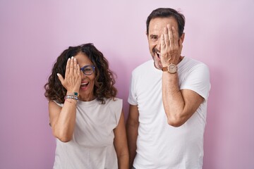 Sticker - Middle age hispanic couple together over pink background covering one eye with hand, confident smile on face and surprise emotion.