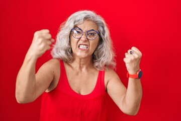 Sticker - Middle age woman with grey hair standing over red background angry and mad raising fists frustrated and furious while shouting with anger. rage and aggressive concept.