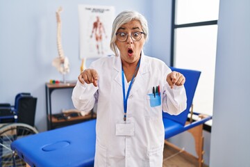 Poster - Middle age woman with grey hair working at pain recovery clinic pointing down with fingers showing advertisement, surprised face and open mouth