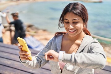 Poster - Young hispanic woman wearing sportswear looking stopwatch using smartphone at seaside