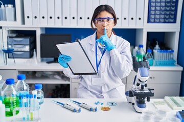 Poster - Hispanic young woman working at scientist laboratory asking to be quiet with finger on lips. silence and secret concept.