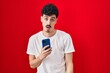 Hispanic man using smartphone over red background in shock face, looking skeptical and sarcastic, surprised with open mouth