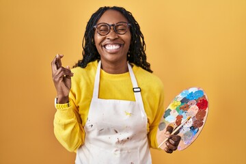 Poster - African woman holding painter palette gesturing finger crossed smiling with hope and eyes closed. luck and superstitious concept.