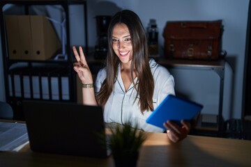 Canvas Print - Young brunette woman working at the office at night smiling looking to the camera showing fingers doing victory sign. number two.