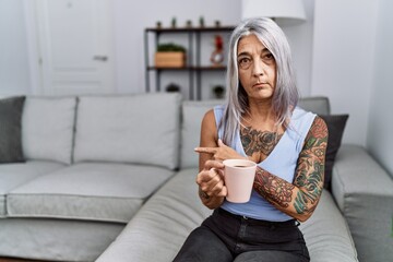 Canvas Print - Middle age grey-haired woman drinking coffee sitting on the sofa at home pointing with hand finger to the side showing advertisement, serious and calm face