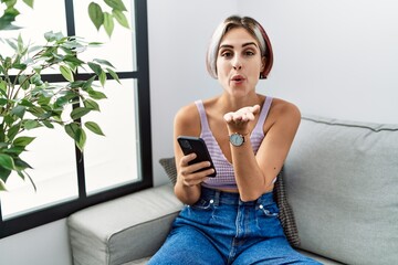 Sticker - Young beautiful woman using smartphone typing message sitting on the sofa looking at the camera blowing a kiss with hand on air being lovely and sexy. love expression.