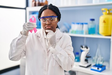 Wall Mural - African american woman working at scientist laboratory holding pink ribbon serious face thinking about question with hand on chin, thoughtful about confusing idea