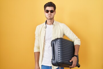 Poster - Young hispanic man holding suitcase going on summer vacation puffing cheeks with funny face. mouth inflated with air, crazy expression.