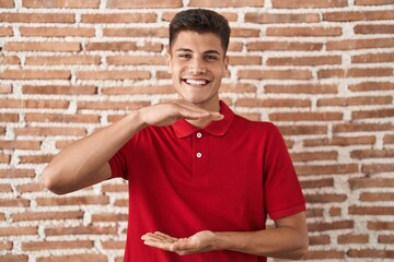 Canvas Print - Young hispanic man standing over bricks wall gesturing with hands showing big and large size sign, measure symbol. smiling looking at the camera. measuring concept.