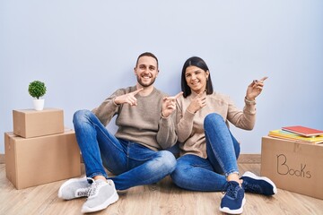 Wall Mural - Young couple moving to a new home smiling and looking at the camera pointing with two hands and fingers to the side.