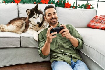 Poster - Young hispanic man playing video game sitting on sofa with dog by christmas decor at home