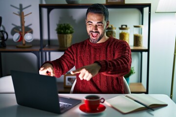 Wall Mural - Young hispanic man with beard using computer laptop at night at home pointing to you and the camera with fingers, smiling positive and cheerful