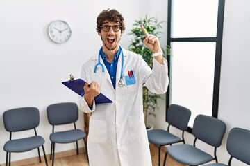 Wall Mural - Young hispanic doctor man at waiting room smiling amazed and surprised and pointing up with fingers and raised arms.