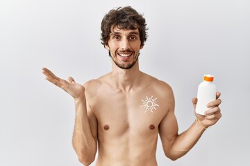 Sticker - Young hispanic man standing shirtless holding sunscreen lotion smiling cheerful presenting and pointing with palm of hand looking at the camera.