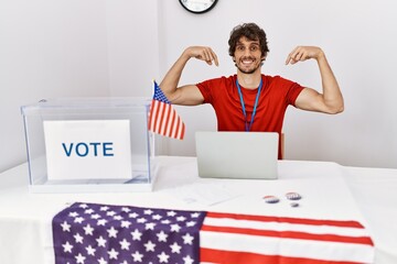 Canvas Print - Young hispanic man at political election sitting by ballot looking confident with smile on face, pointing oneself with fingers proud and happy.