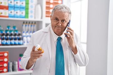 Poster - Middle age grey-haired man pharmacist holding pills bottle talking on smartphone at pharmacy