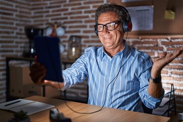 Poster - Middle age hispanic man using touchpad sitting on the table at night smiling cheerful presenting and pointing with palm of hand looking at the camera.