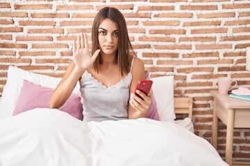 Poster - Young hispanic woman using smartphone sitting on the bed at home with open hand doing stop sign with serious and confident expression, defense gesture