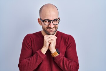 Wall Mural - Young bald man with beard standing over white background wearing glasses laughing nervous and excited with hands on chin looking to the side