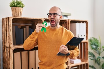 Wall Mural - Young bald man with beard holding support green ribbon angry and mad screaming frustrated and furious, shouting with anger. rage and aggressive concept.