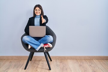 Wall Mural - Young hispanic woman sitting on chair using computer laptop smiling friendly offering handshake as greeting and welcoming. successful business.