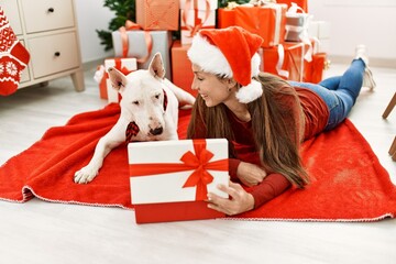 Wall Mural - Young caucasian woman unboxing gift lying with dog by christmas tree at home