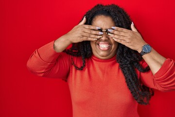 Poster - Plus size hispanic woman standing over red background covering eyes with hands smiling cheerful and funny. blind concept.