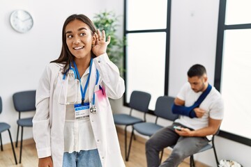 Poster - Young asian doctor woman at waiting room with a man with a broken arm smiling with hand over ear listening an hearing to rumor or gossip. deafness concept.